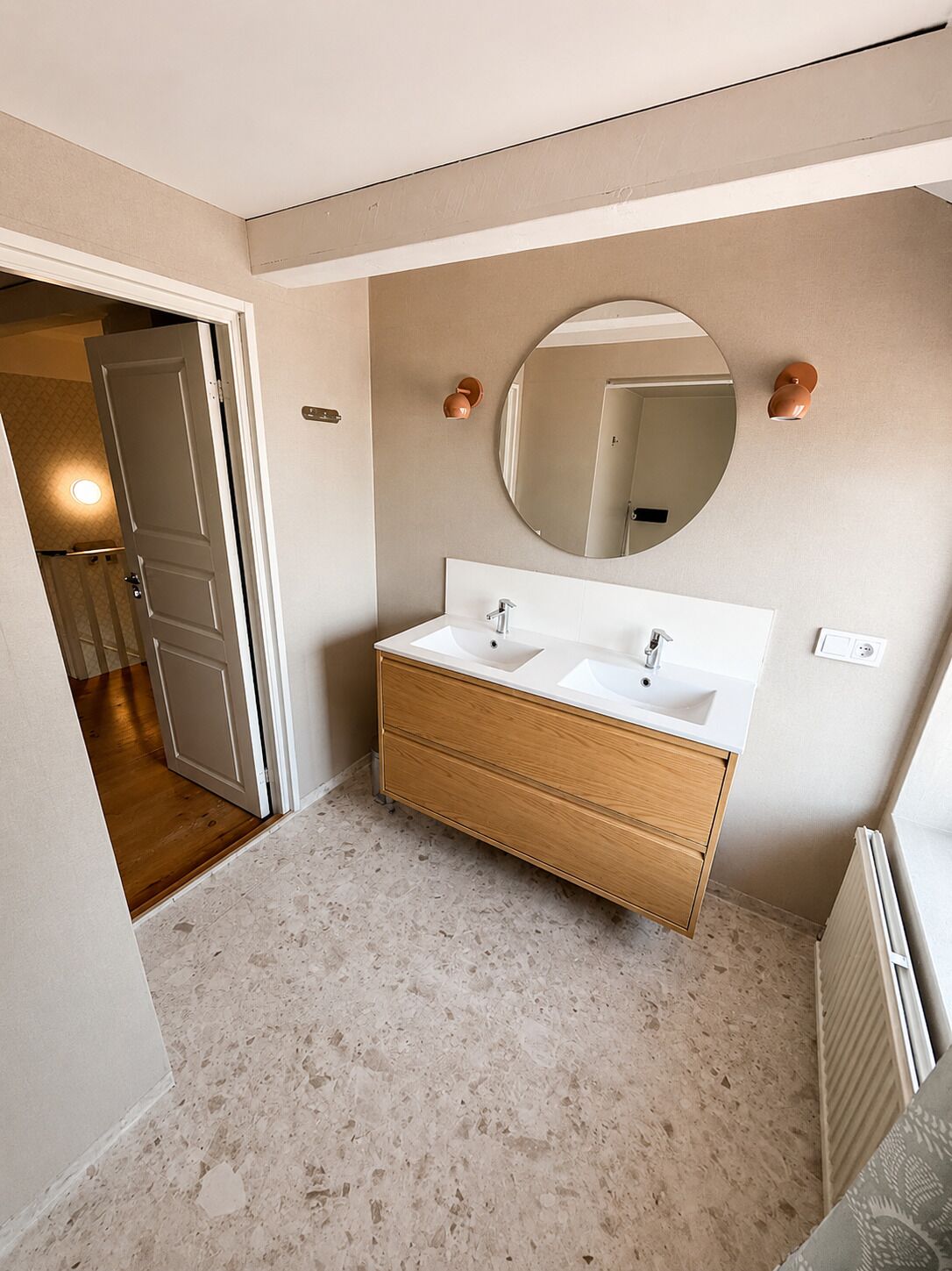 A bathroom with a double sink vanity, a round mirror, and a door leading to another room.
