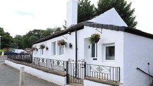 A white building with a black metal fence, windows with hanging flower baskets, and a white door. - White Bungalow With 75" Tv, Air Con, Contractors (Bradford)