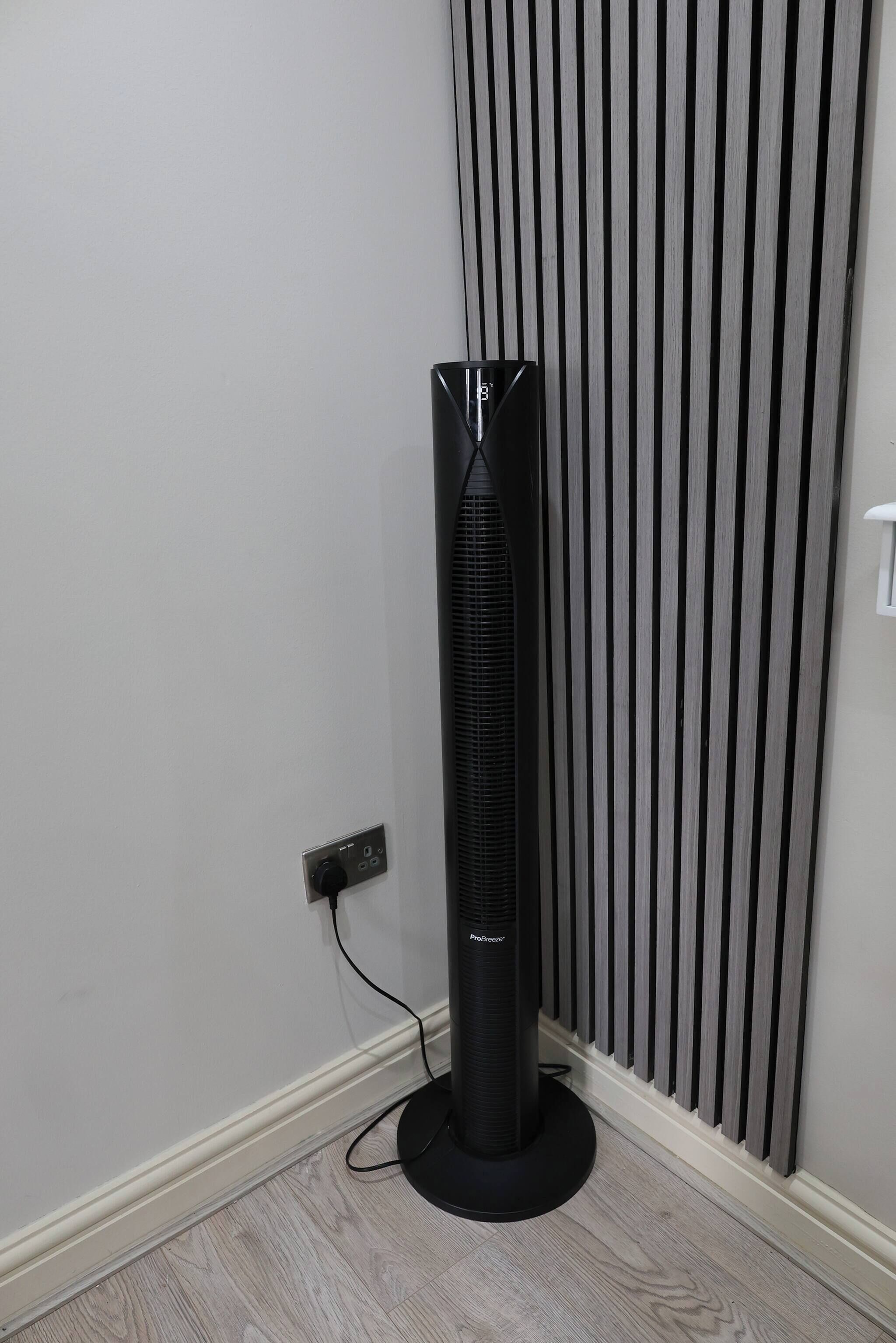 A standing fan in a corner of a room with a white wall and a vertical striped panel.