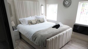 A bedroom with a bed, a bedside table, a lamp, a clock, and a window. - White Bungalow With 75" Tv, Air Con, Contractors (Bradford)