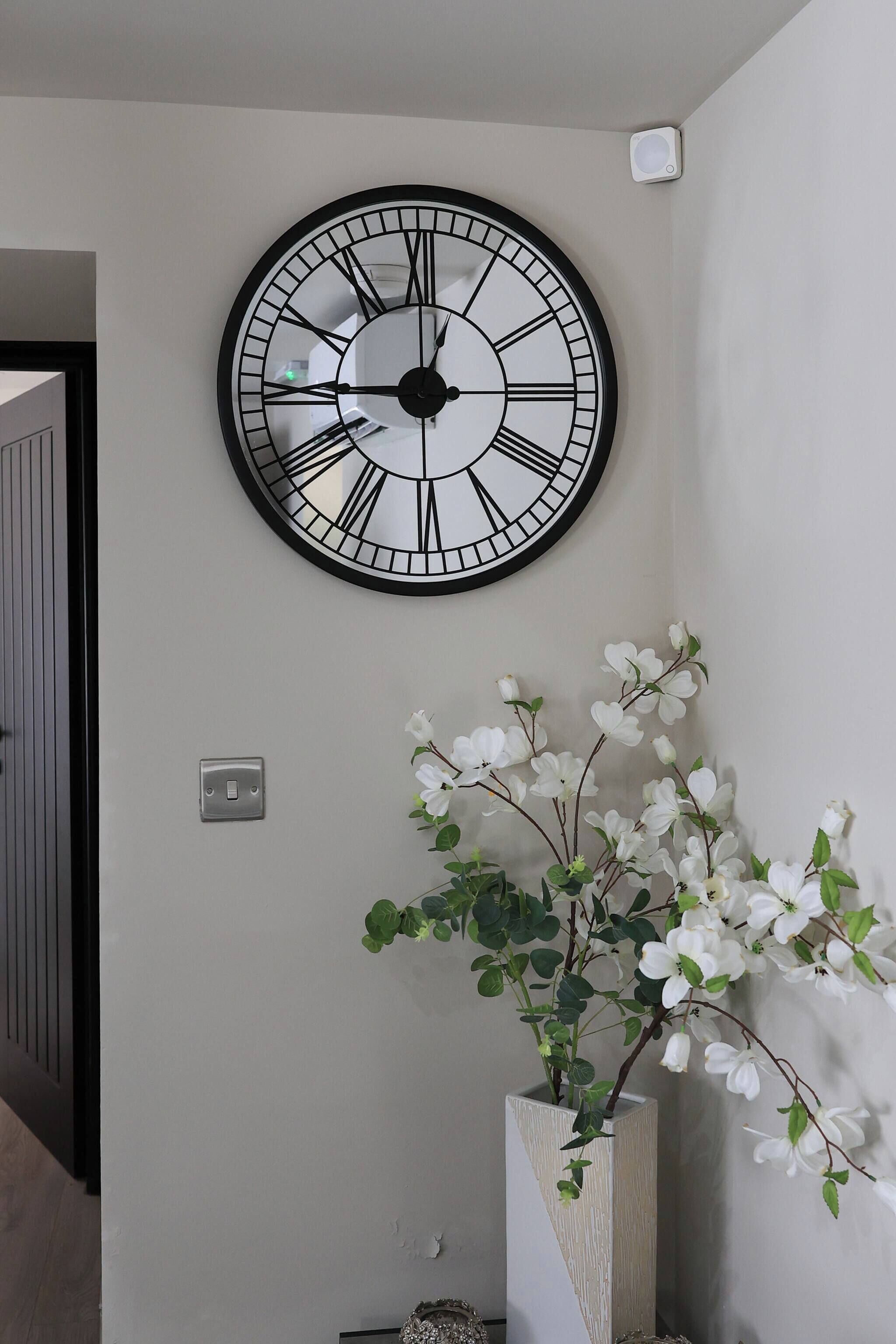 A wall-mounted clock with Roman numerals, a light switch, and a white vase with artificial flowers.