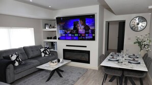 A modern living room with a flat-screen TV, a fireplace, a dining area with a table set, and a clock on the wall. - White Bungalow With 75" Tv, Air Con, Contractors (Bradford)