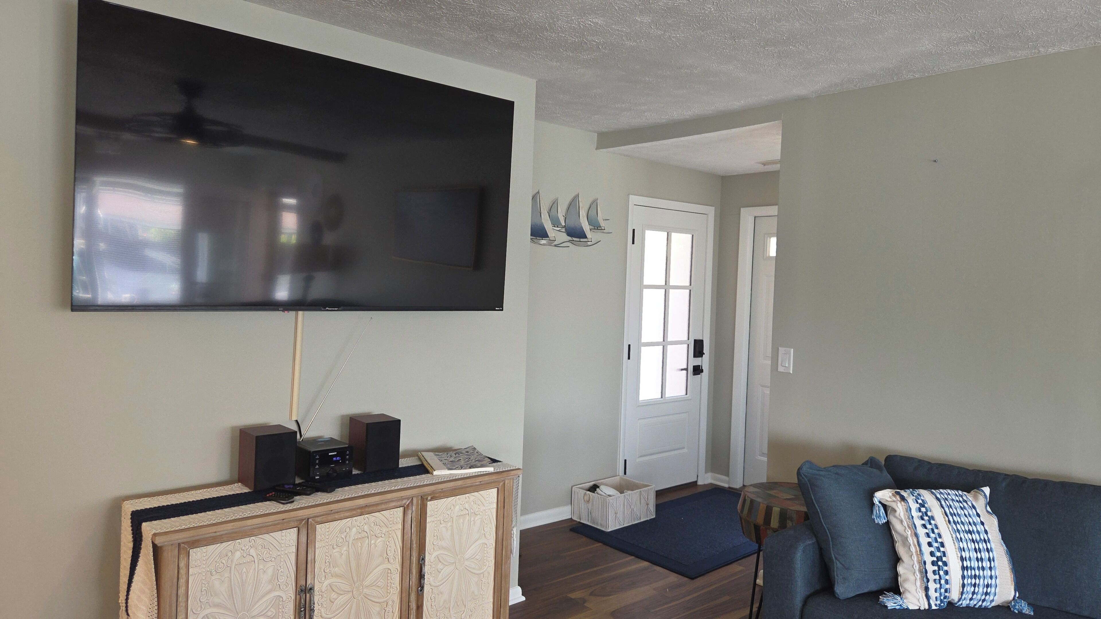A living room with a flat-screen TV mounted on the wall, a wooden console table with speakers, a blue sofa, and a door leading to another room.