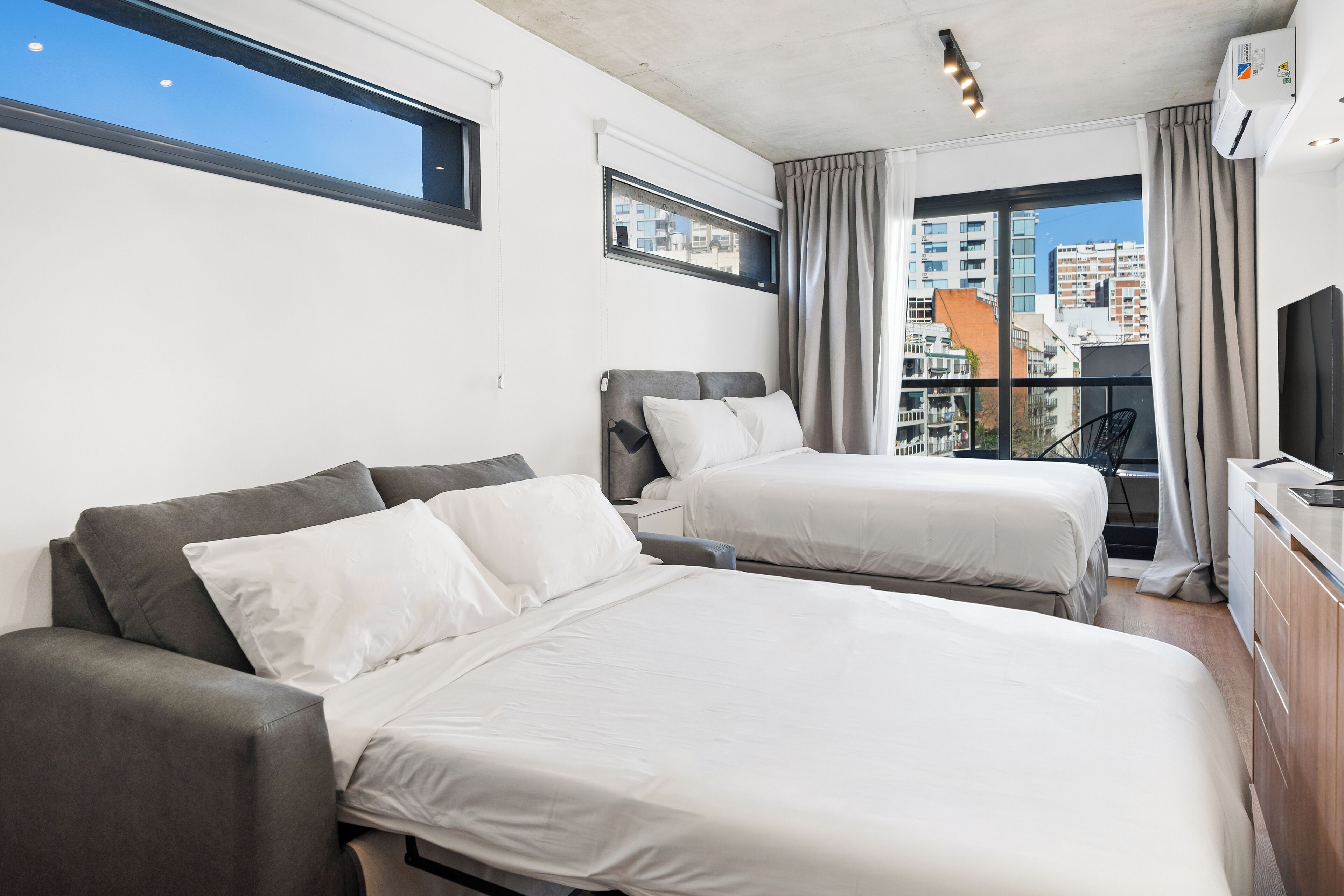 Habitación de hotel con dos camas, televisión, balcón con vistas a edificios y ventana con cortinas.