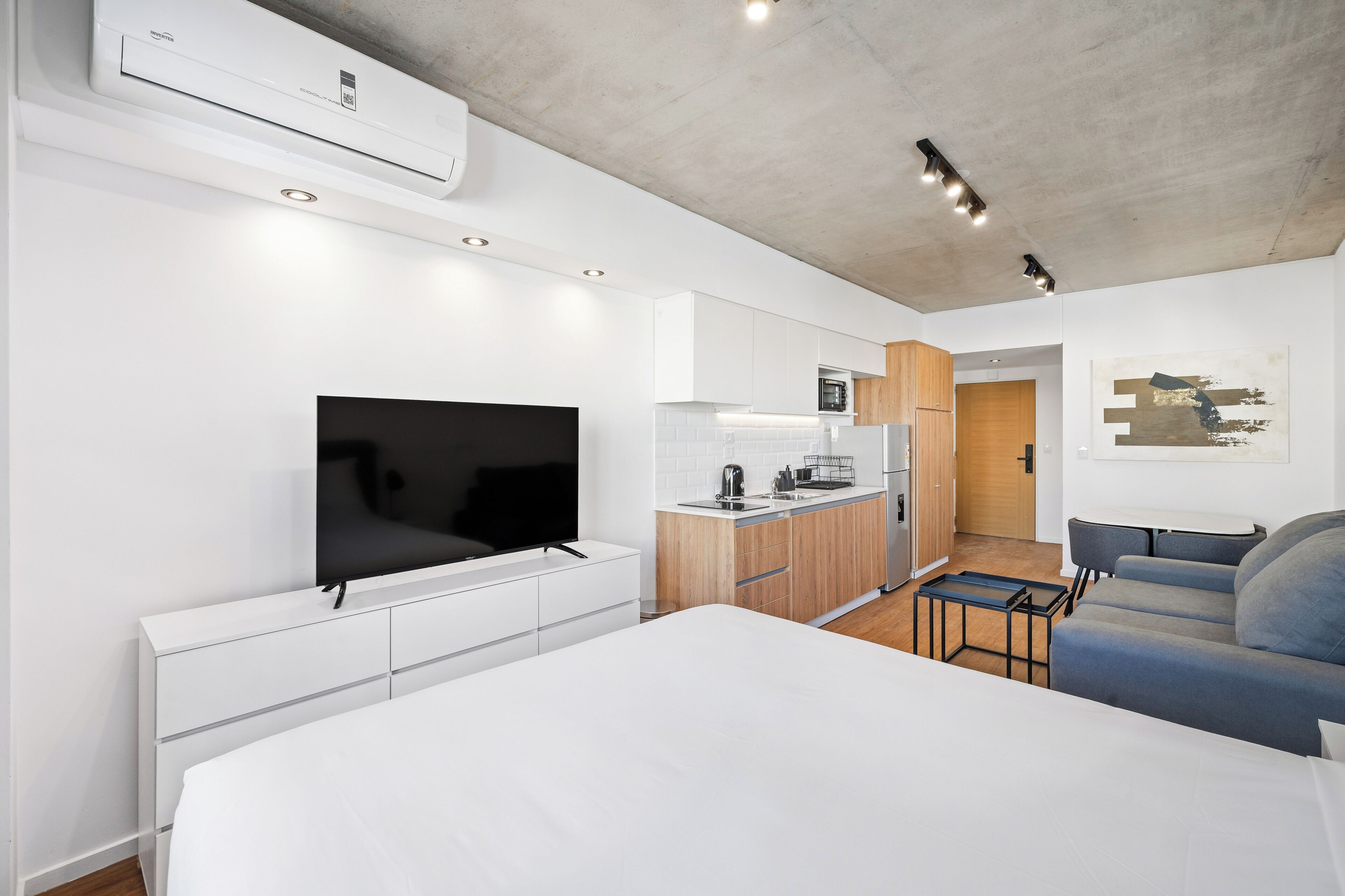 A modern hotel room with a bed, a flat-screen TV, a kitchenette, and a seating area.