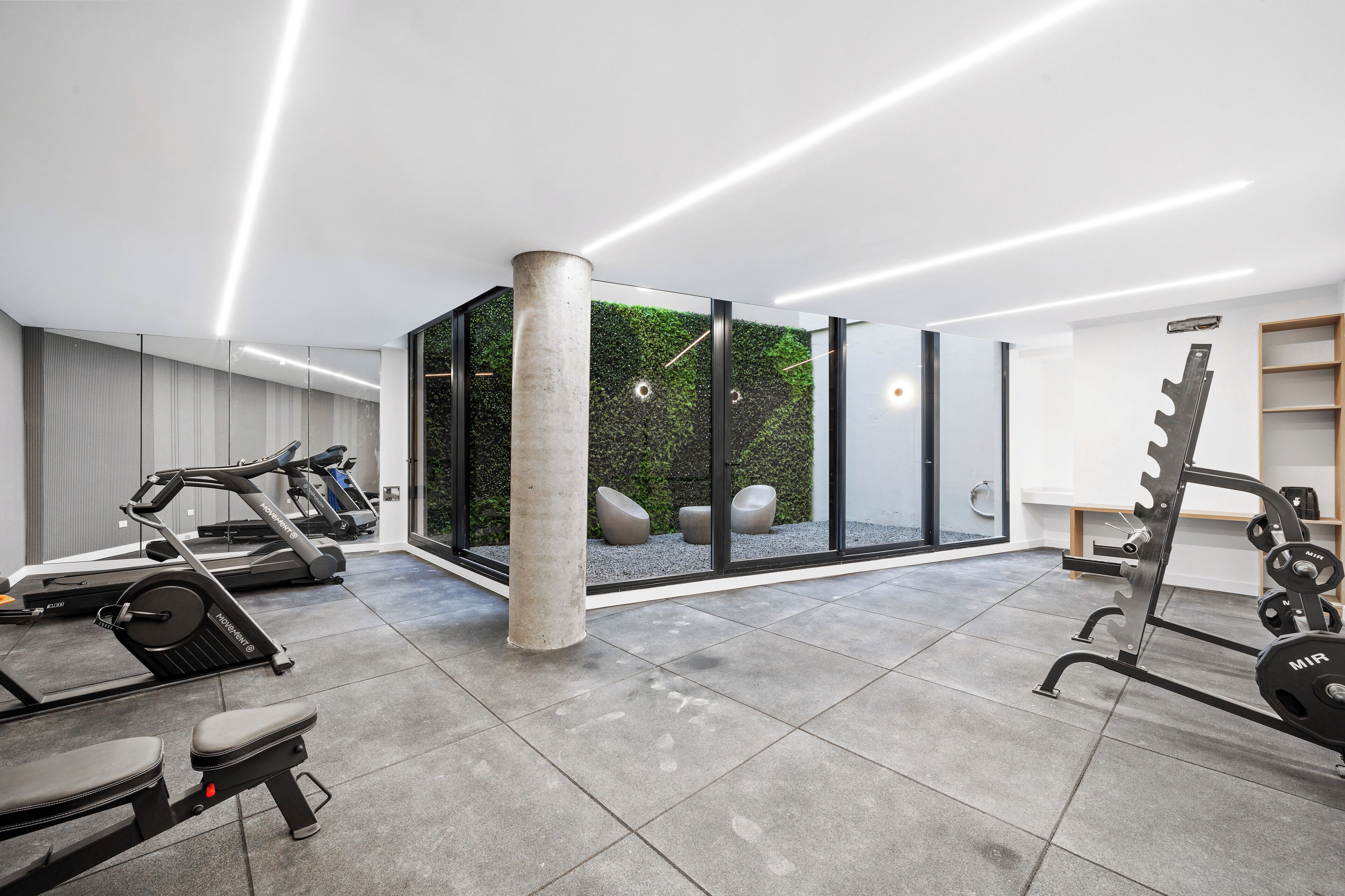 A modern gym with exercise equipment, a concrete pillar, and a glass wall with a view of greenery.