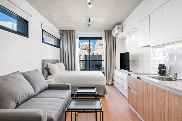 A compact hotel room with a sofa, a bed, a small table, a TV, and a kitchenette.