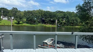Una terraza de madera con vistas a un lago, un barco amarrado en el muelle y una estructura de piedra al fondo.