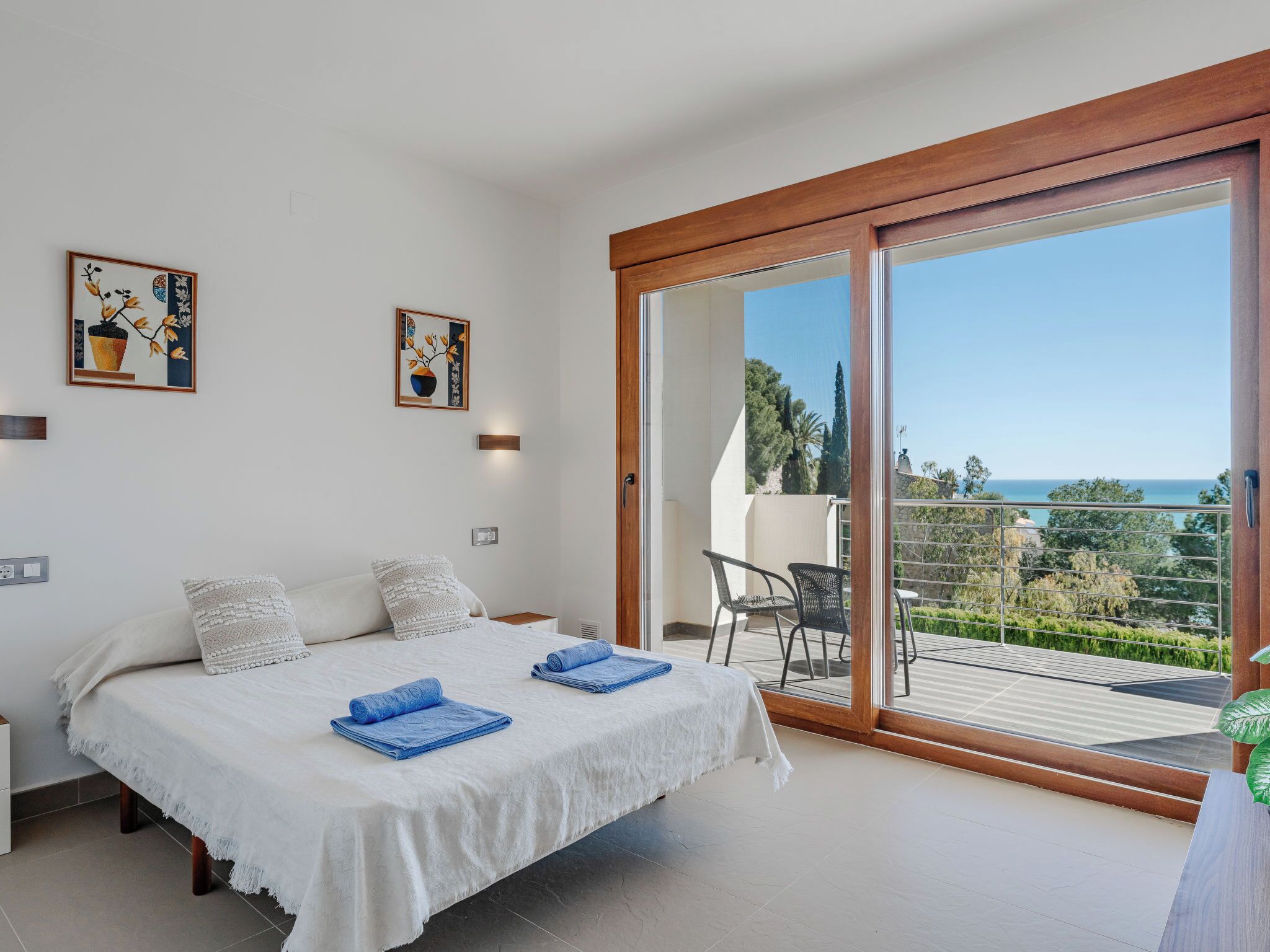 A bedroom with a bed, two pillows, a nightstand, a wall-mounted light, and a large sliding glass door leading to an outdoor balcony with a view of the sea.