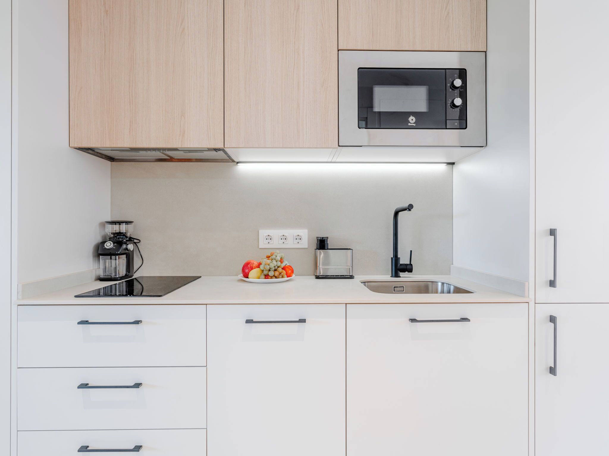 Een moderne keuken met witte kasten, een ingebouwde magnetron, een koffiezetapparaat, een fruitschaal, een glazen pot en een spoelbak met een zwarte kraan.