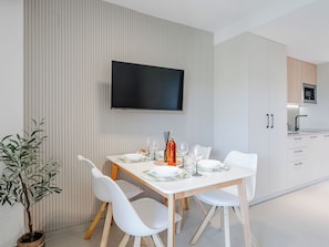 A modern dining area with a white table, white chairs, a flat-screen TV mounted on the wall, and a kitchen with white cabinets and a microwave. - Playa Sur by Interhome (Dénia)