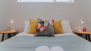 Una cama con un cojín floral, dos mesitas de noche con lámparas y una ventana arriba.