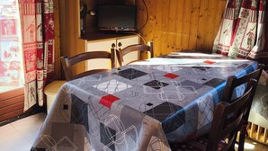 Een eetruimte met een tafel, bedekt met een tafelkleed met een geometrisch patroon, houten stoelen, een kast met glazen deuren, een klok en een ingelijst kunstwerk aan de muur.