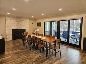 Un vaste espace dédié à la salle à manger, avec une longue table et des chaises en bois, une cheminée, et une vue sur une terrasse extérieure accessible par de grandes portes coulissantes en verre.