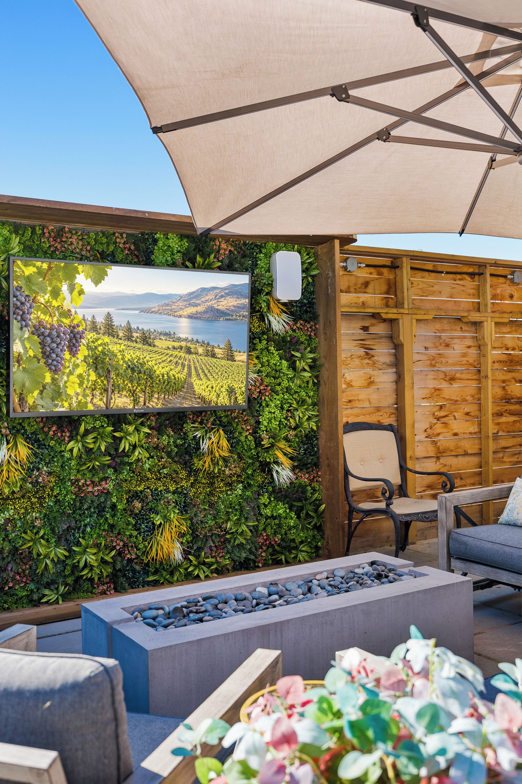 Eine Terrasse mit flachbildschirmfernseher an der Wand, umgeben von üppiger Bepflanzung und einer Sitzgruppe mit Bank und Tisch.