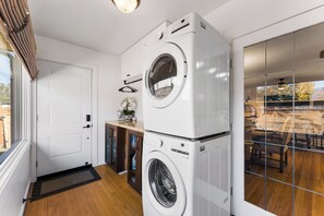 A laundry room with two stacked front-loading washing machines, a door, a small cabinet, and a window. - Luxury French-Inspired Retreat • Private Terrace (Kelowna)