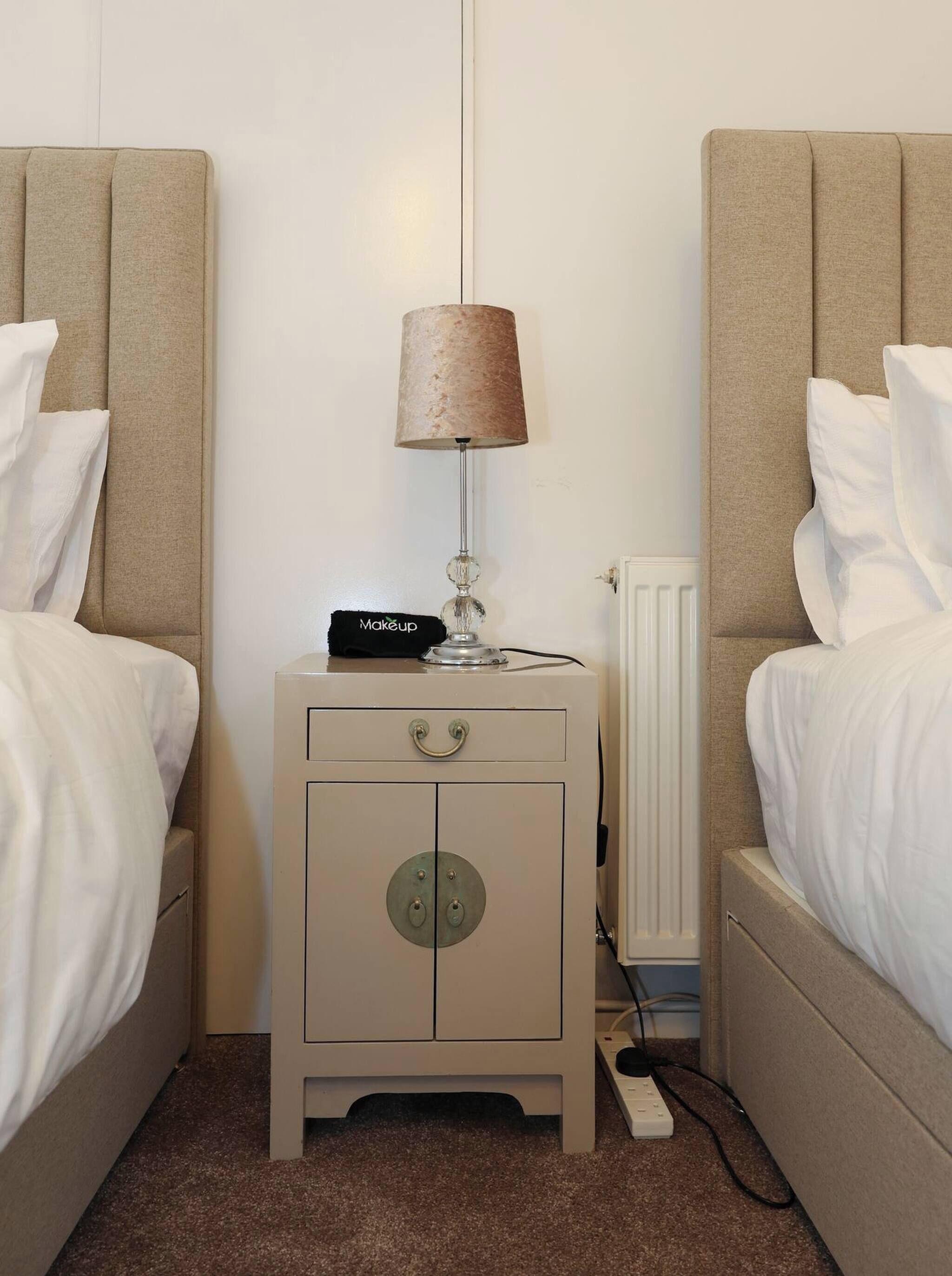 A small, neutral-colored room with two beds, a bedside table with a lamp, a small cabinet, and a radiator.