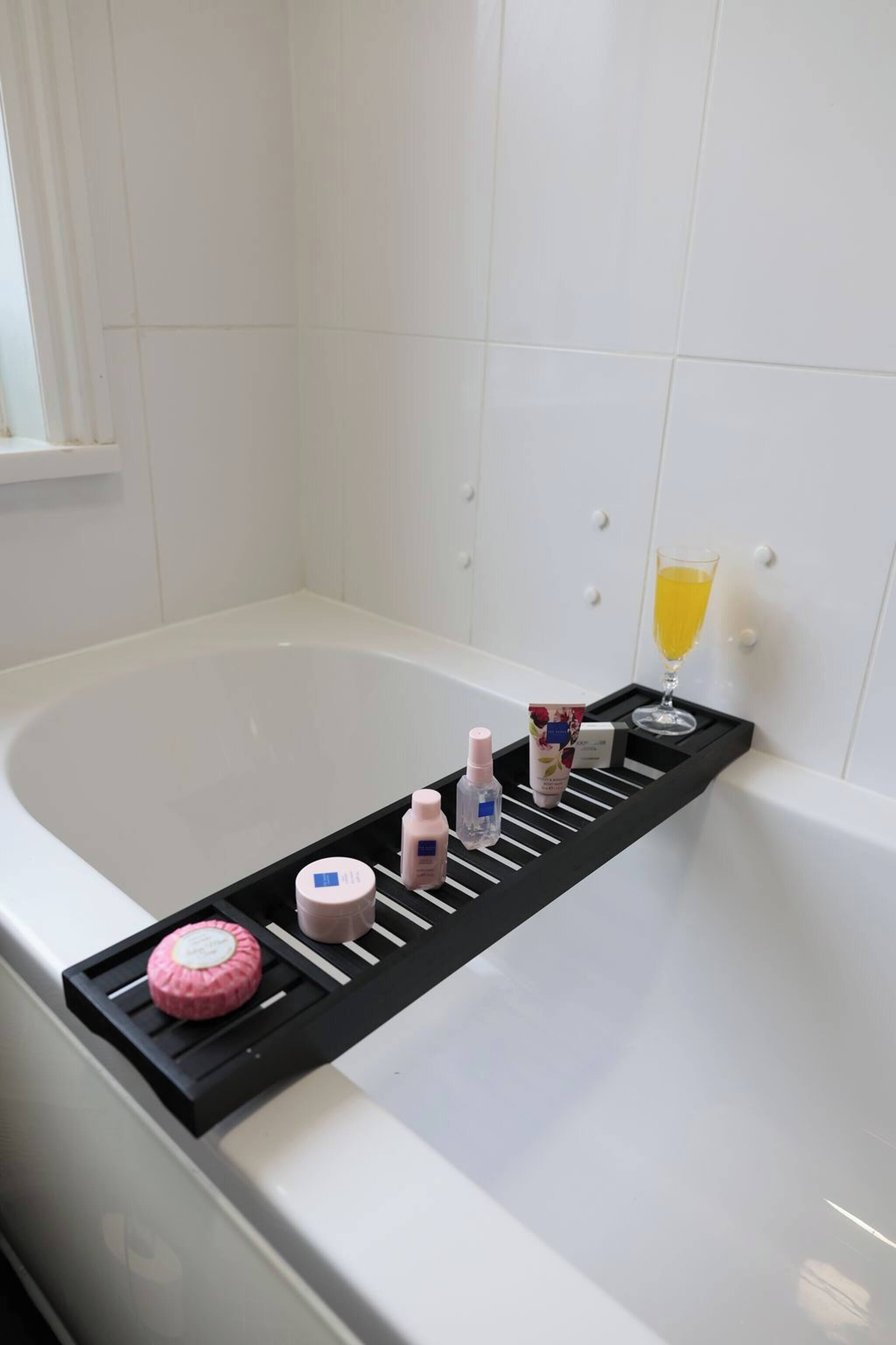 A bathtub with a black tray holding toiletries, a glass of yellow liquid, and a pink object.