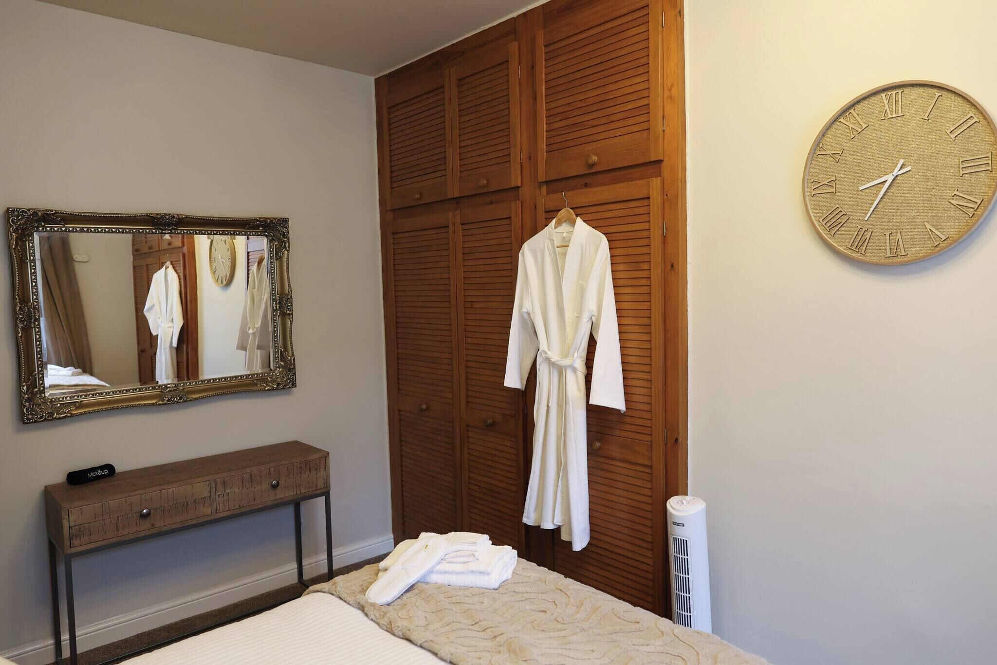 A bedroom with a wooden wardrobe, a mirror, a bedside table, a clock, and a robe hanging on the wall.