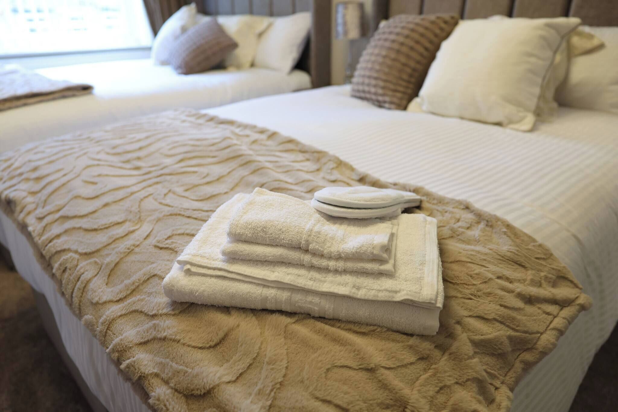 Una cama bien hecha con una manta beige texturizada, una toalla blanca doblada y una pequeña bandeja blanca sobre la cama.