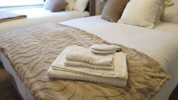 Una cama bien hecha con una manta beige texturizada, una toalla blanca doblada y una pequeña bandeja blanca sobre la cama.