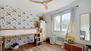 Una cameretta con un letto a castello, una ventola a soffitto, una finestra con tende, un termosifone e un piccolo armadietto pieno di giocattoli.