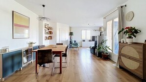 Un'ampia zona giorno con un tavolo da pranzo in legno, sedie, un divano e una TV. Si notano mensole con libri e oggetti decorativi, e un orologio a parete.