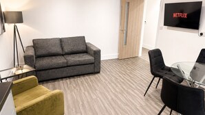 Una sala de estar moderna con un sofá gris, un sillón amarillo, una mesa de centro de vidrio, sillas negras y un televisor que muestra 'NETFLIX'.