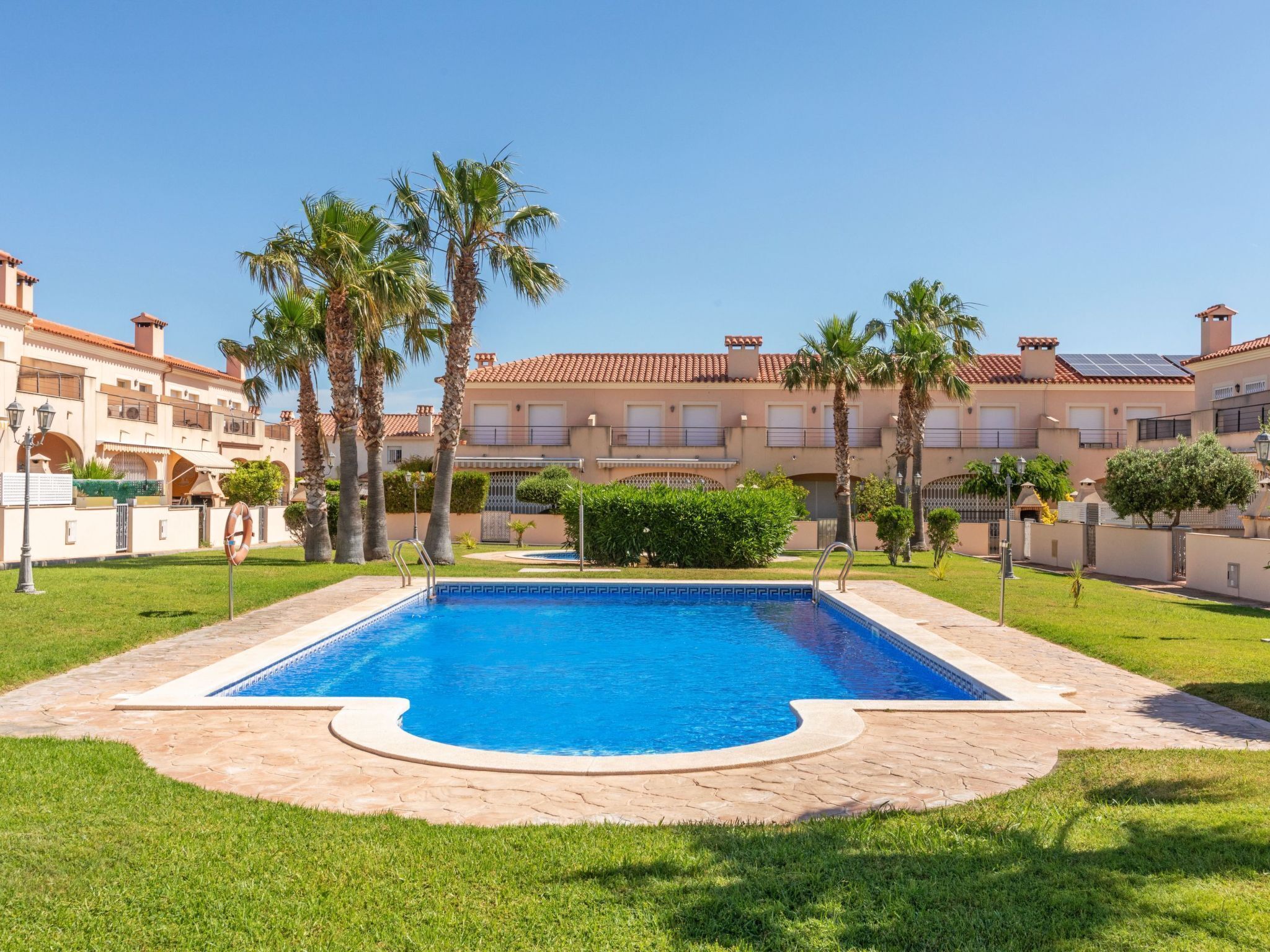 A swimming pool with a tiled deck, surrounded by palm trees and a building with a tiled roof.