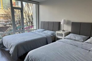 A hotel room with two beds, a nightstand, a lamp, and a window with a view of buildings and bare trees.