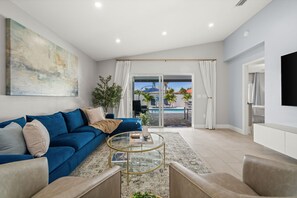 Een moderne woonkamer met een blauwe bank, een glazen salontafel en uitzicht op een zwembad door glazen deuren.