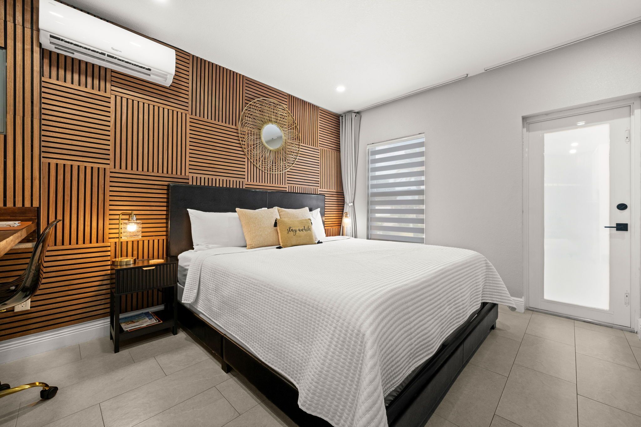 A modern bedroom with a bed, bedside tables, a wall-mounted air conditioner, and a window with blinds.