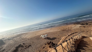 Une plage avec un escalier en bois menant à la grève, une zone sablonneuse parsemée d’empreintes et l’océan en arrière-plan.