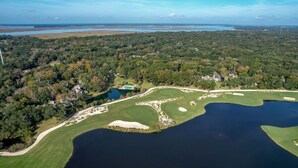 Vista aérea de un campo de golf con un lago, rodeado de un bosque denso y algunos edificios.