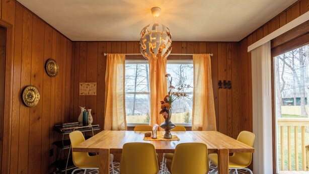 A dining room with a wooden table, yellow chairs, a chandelier, and a window with curtains.