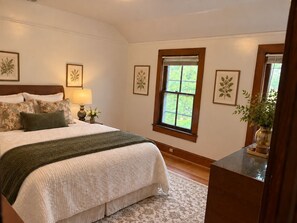 A bedroom with a large bed, a nightstand with a lamp, framed botanical prints on the walls, and a window with a view of greenery.