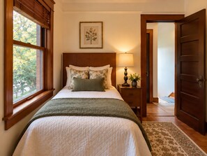 A bedroom with a bed, a nightstand, a lamp, a framed picture on the wall, and a window with blinds.