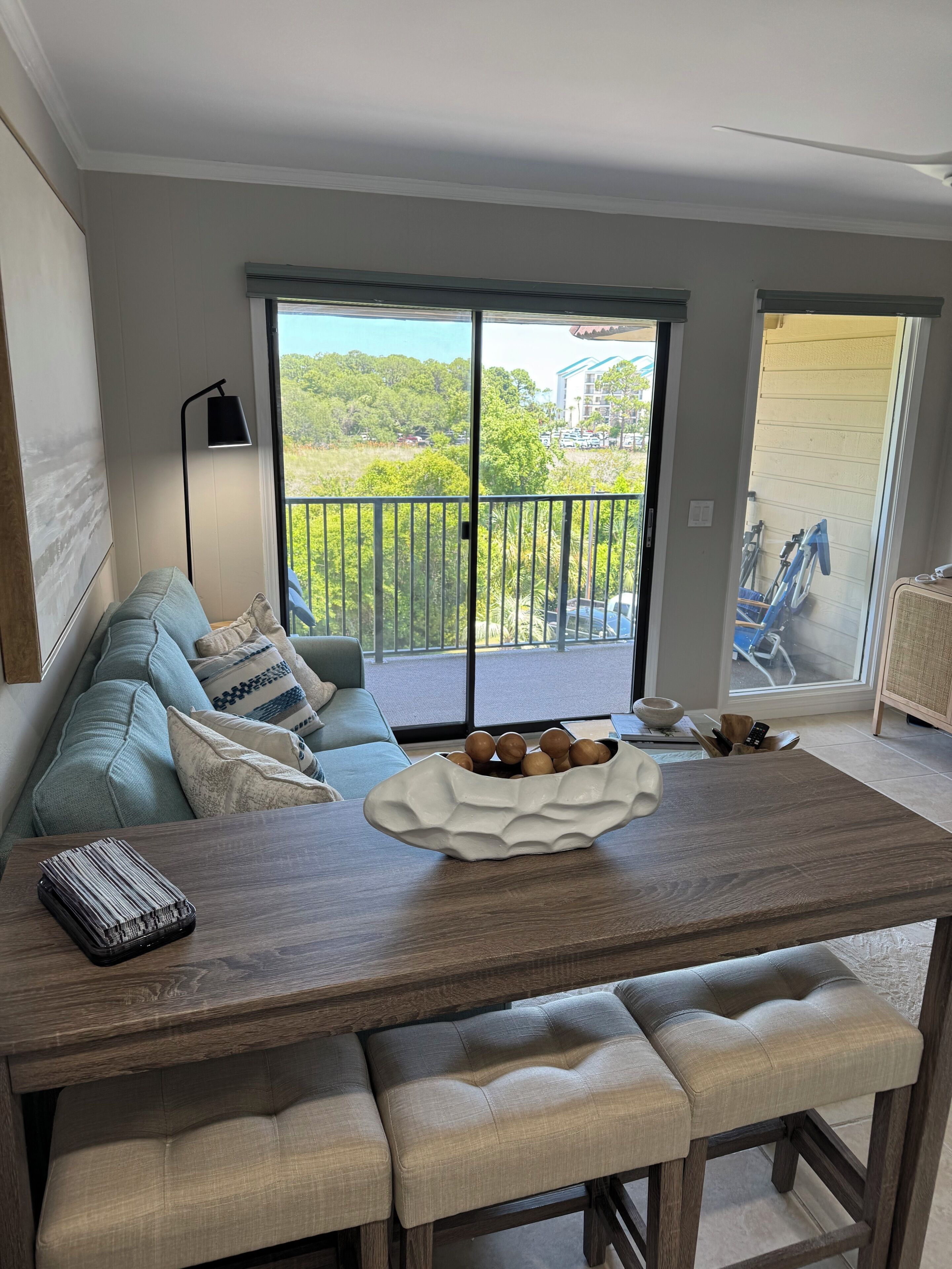 A dining area with a wooden table, cushioned benches, a bowl of fruit, a sofa with cushions, a floor lamp, and a view through a sliding glass door.