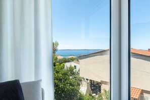 A room with a large window overlooking a coastal view, a bed with a gray and white patterned bedspread, and a window blind partially drawn. - Villa Galeb - Apartment with Private Pool and Sea View (Petrčane (Zadar))