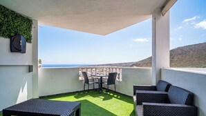 Un espacio de estar al aire libre techado con mesa y sillas, piso de césped artificial y vistas al mar y las colinas.
