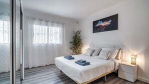 Un dormitorio con una cama, mesitas de noche, una ventana con cortinas, una planta y un cuadro en la pared.