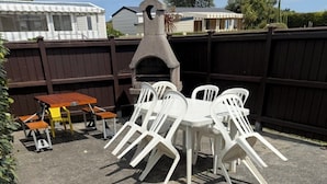 A patio with a round table and white plastic chairs, a stone oven, and a wooden table with chairs.