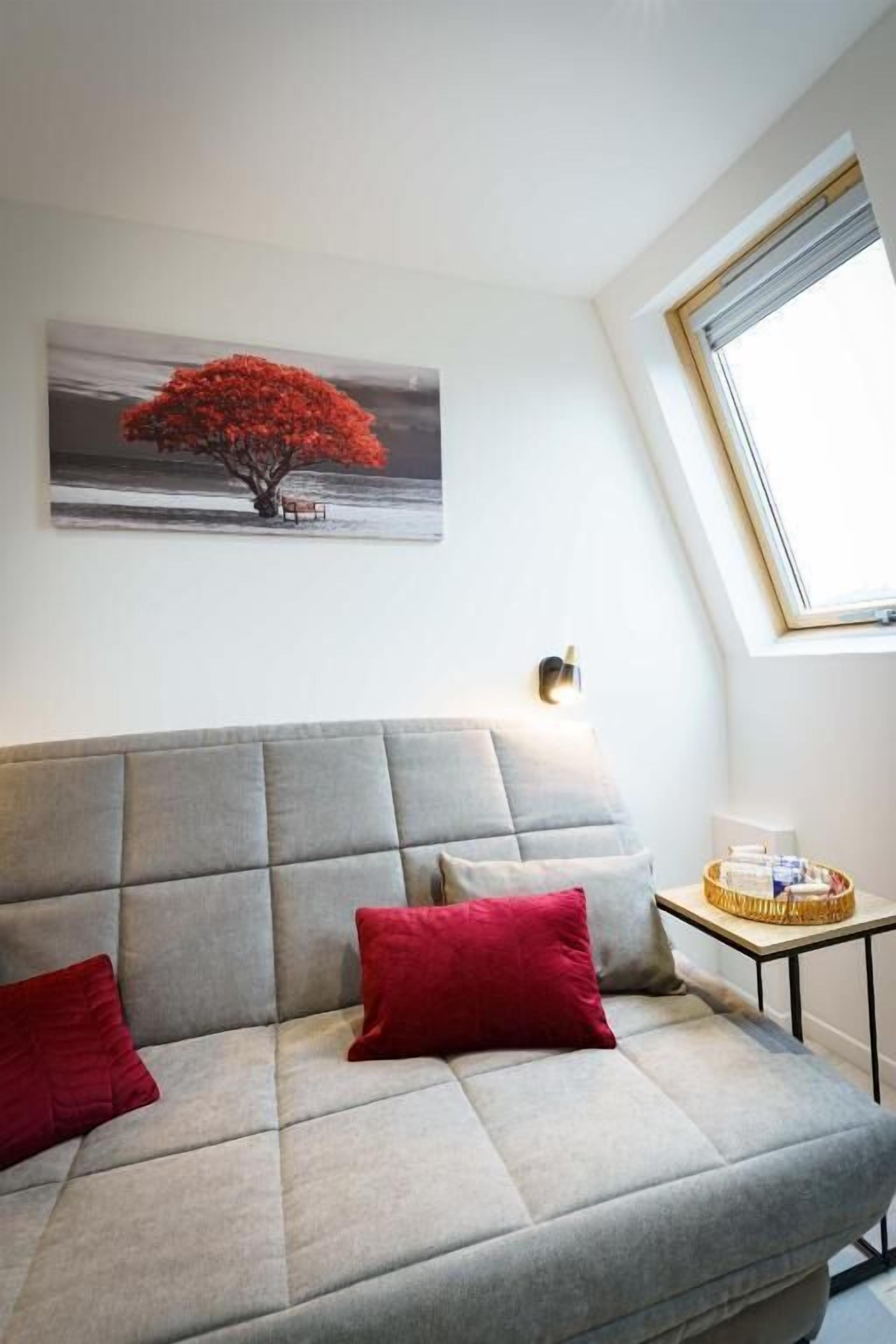 A modern living room with a grey sofa, red cushions, a wall-mounted artwork of a tree, and a small table with a basket.