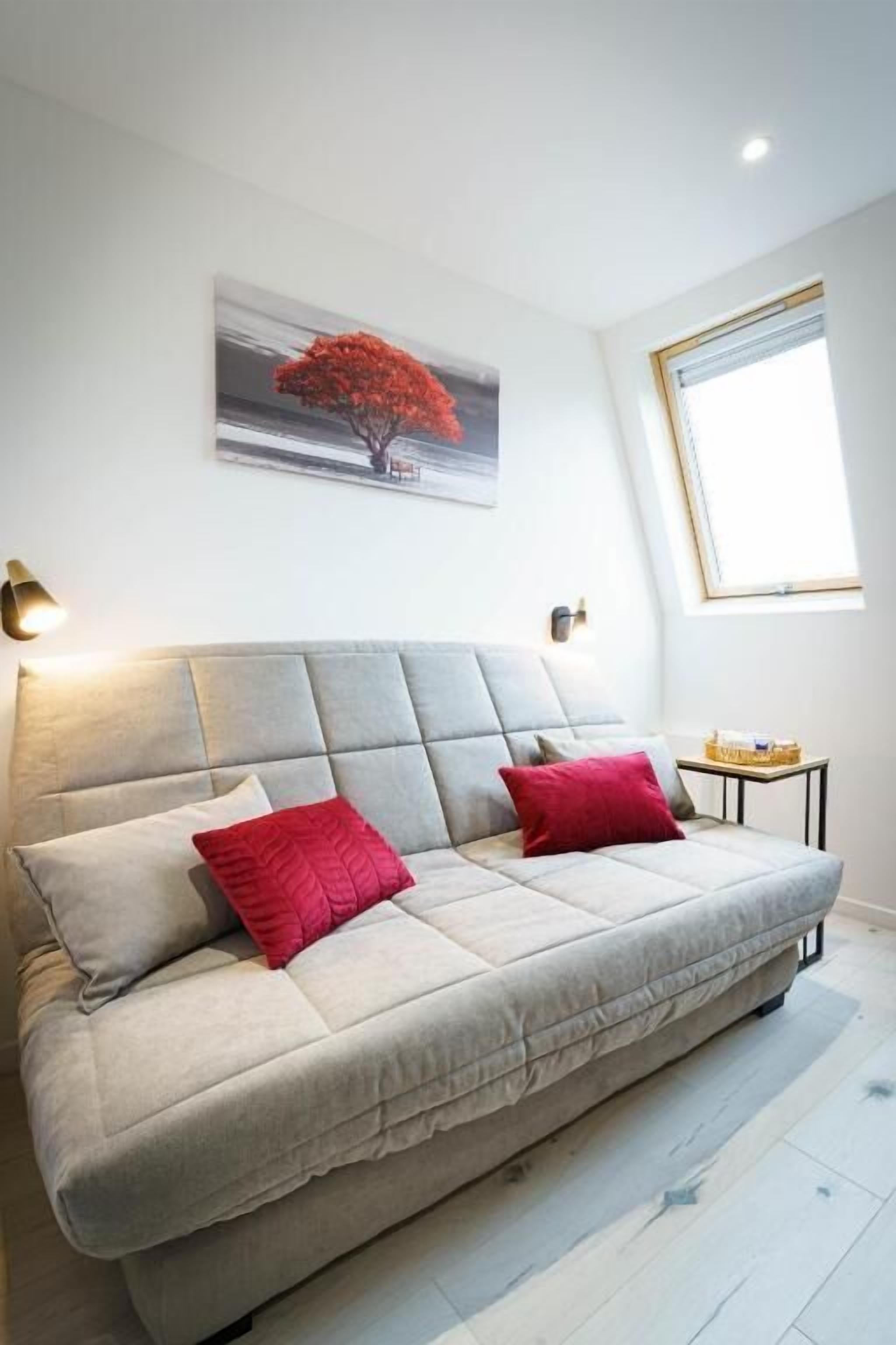 A room with a sofa, two red pillows, a painting of a tree, a window, and a small table with a lamp.