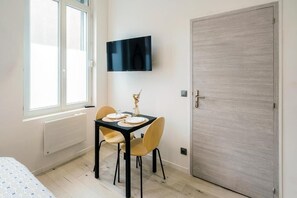 Quarto com cama, mesa e cadeiras, televisão na parede e uma porta.