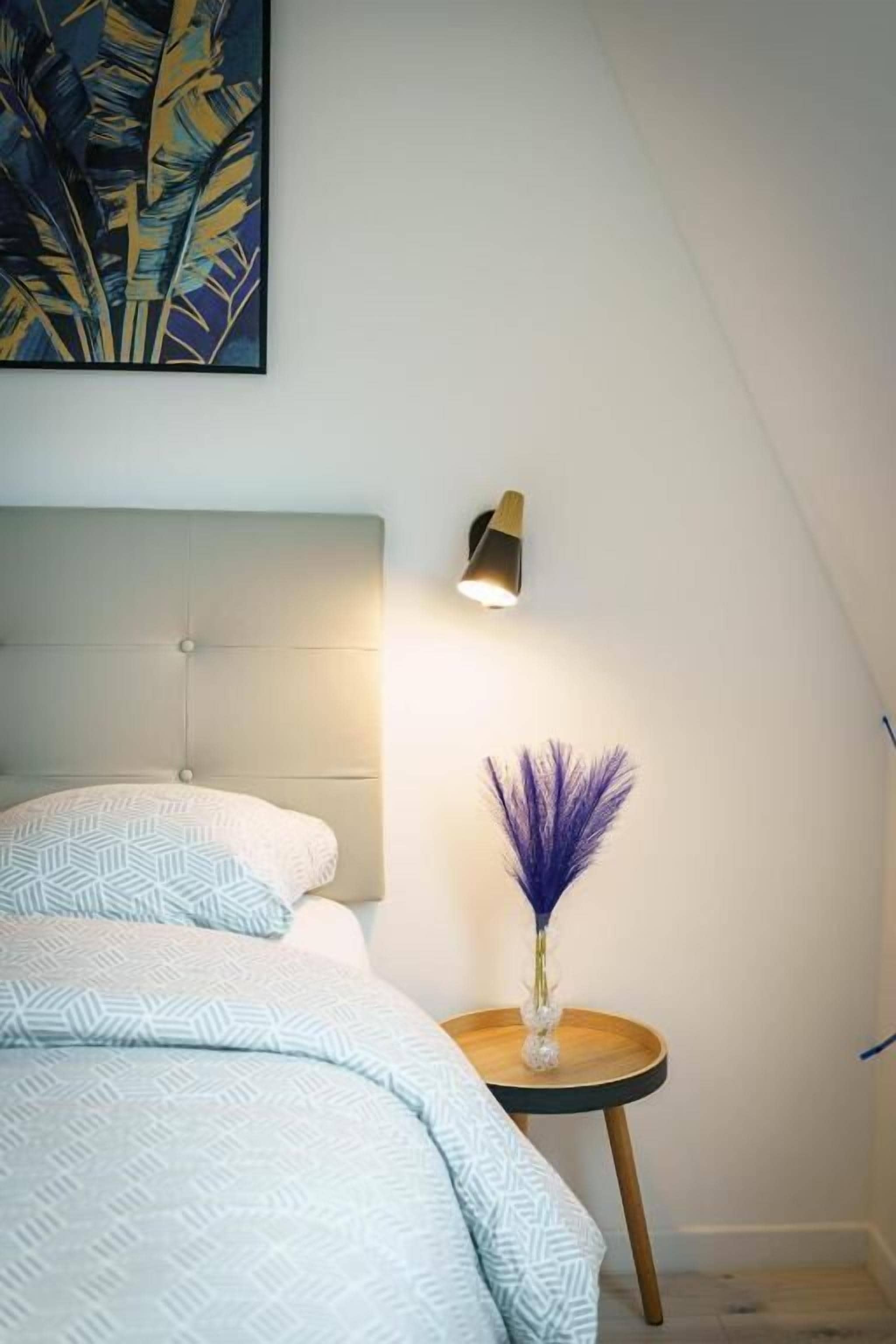 Quarto com cama, mesa de cabeceira com vaso, luminária de parede e um quadro.
