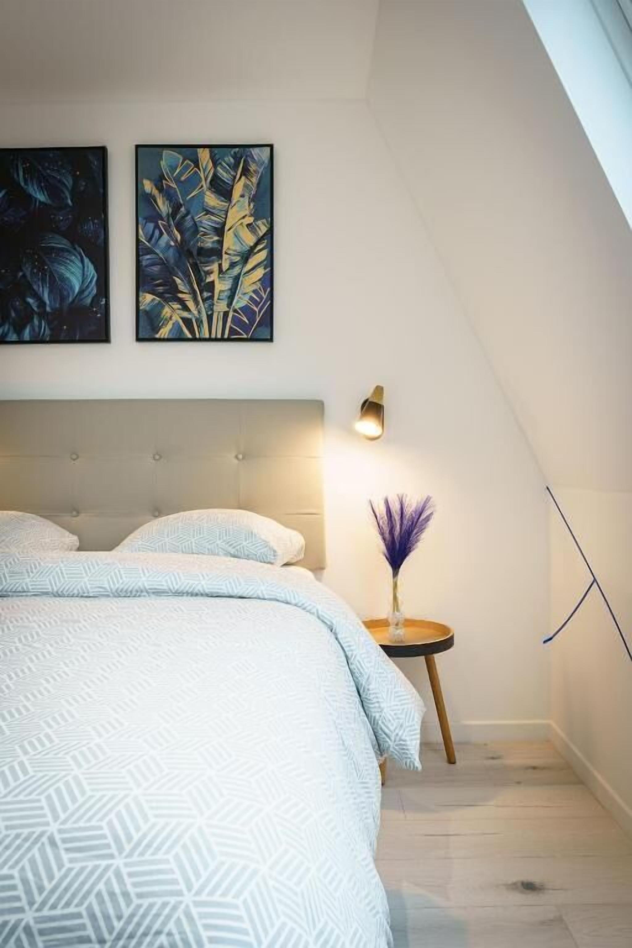 A bedroom with a bed, a bedside table with a lamp, and two wall-mounted artworks.