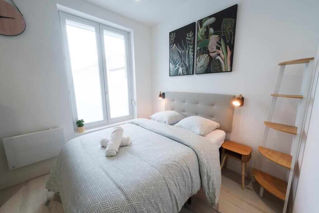 A bedroom with a bed, a window, a radiator, a bedside table, a wall-mounted shelf, and a painting.