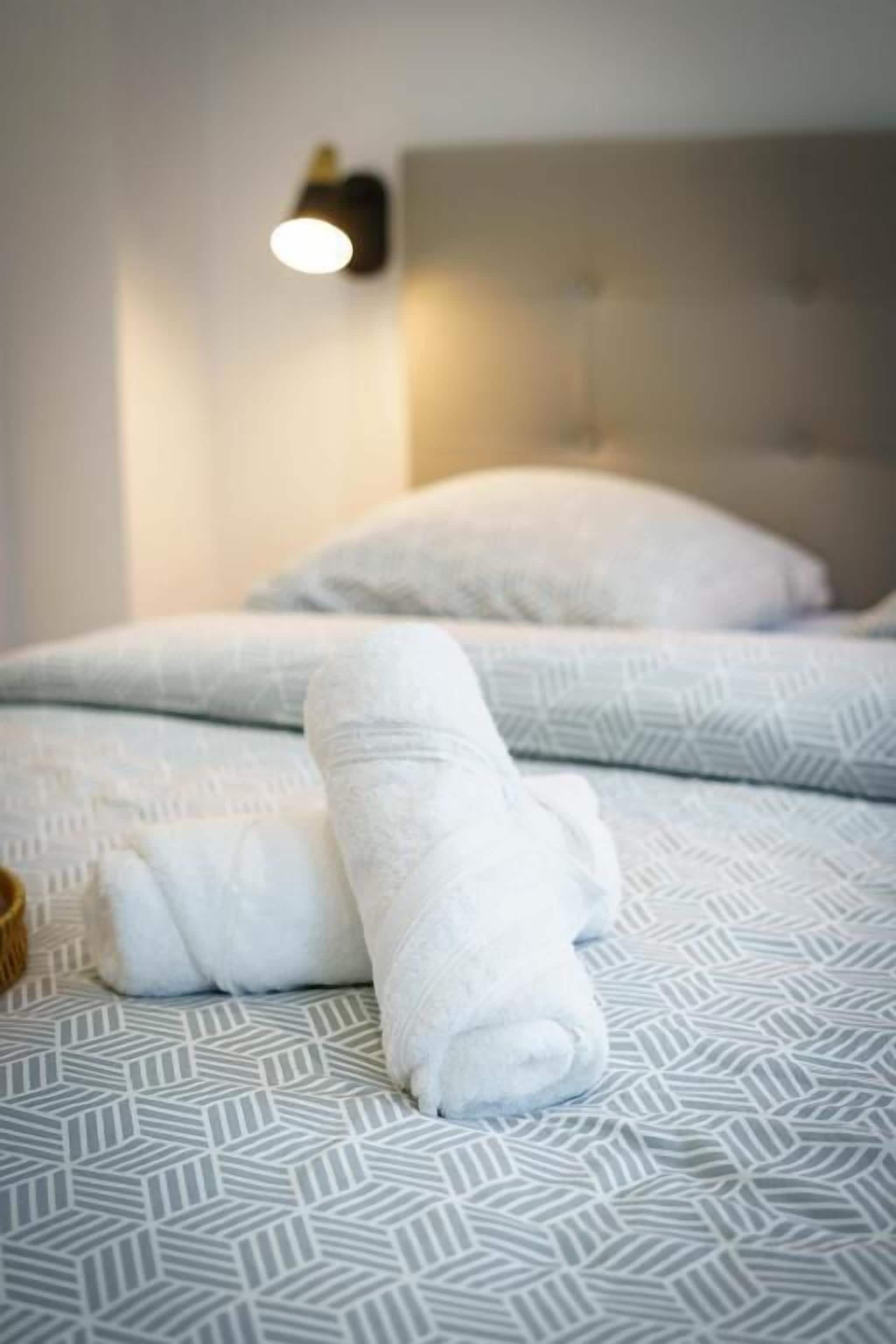 A bed with a patterned bedspread, two rolled towels, and a wall-mounted light fixture.