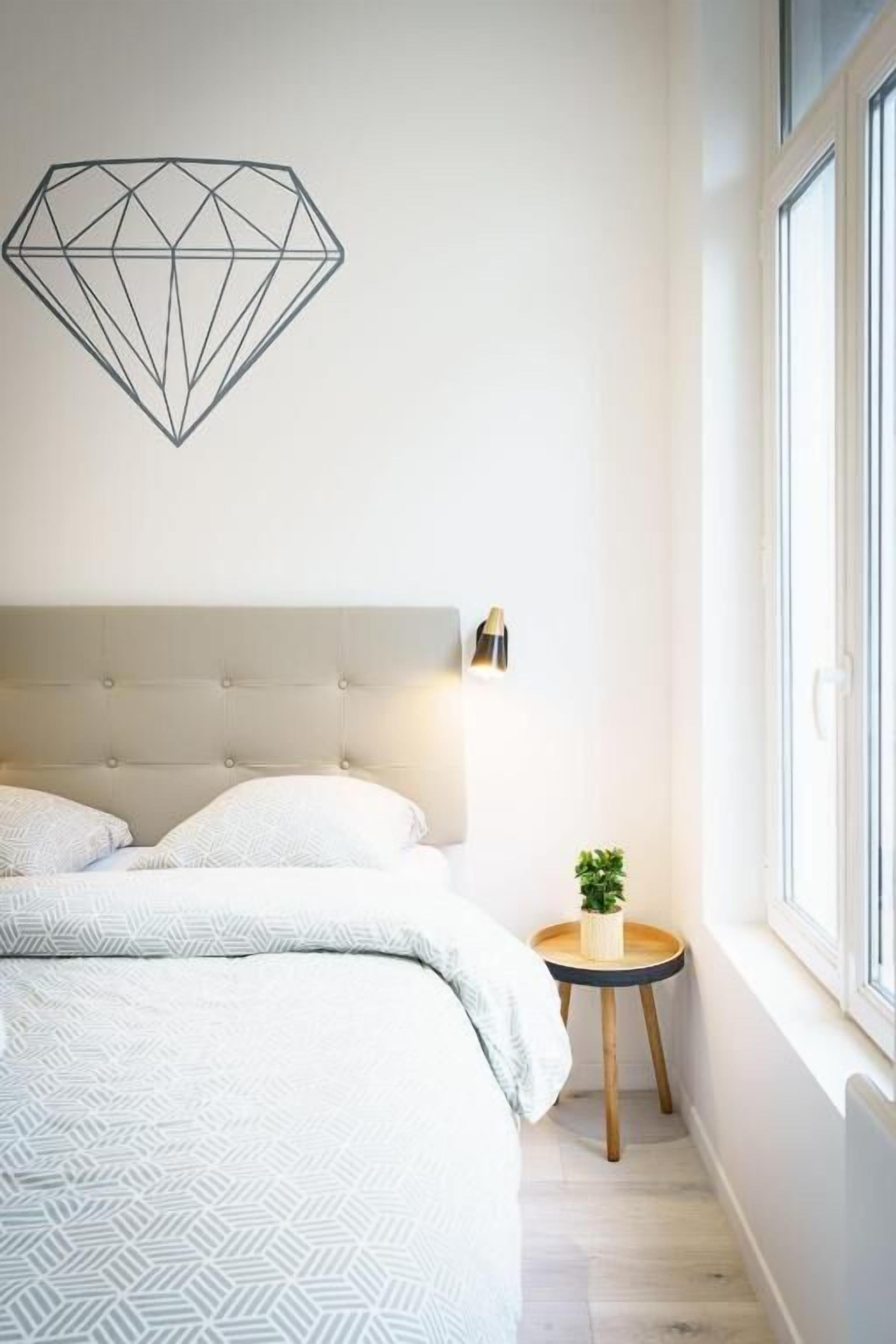 Ein Schlafzimmer mit einem Bett, einer geometrischen Wandleuchte, einem Nachttisch mit Pflanze und einem Fenster.